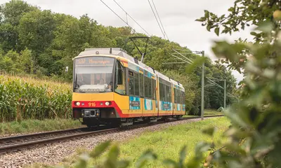 Eine AVG-Stadtbahn fährt auf einem Streckenabschnitt der Kraichtalbahn. Im Hintergrund sind bewaldete Hügel zu sehen.