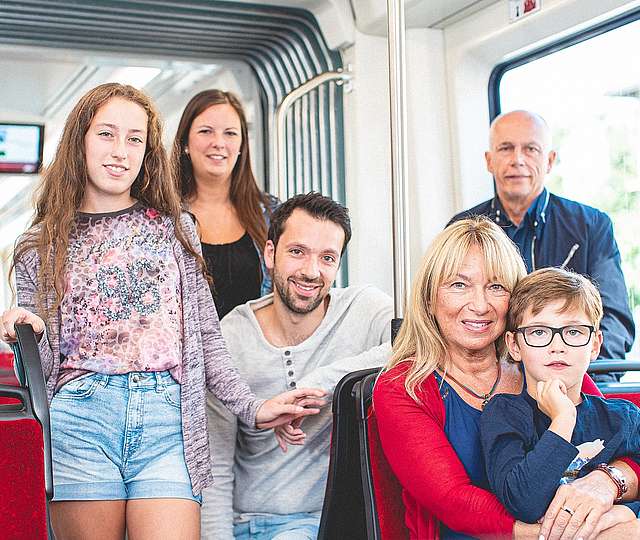 Eine Familie in der Bahn. Links ein brunettes Mädchen mit einer kurzen Jeans Shorts. Im Hintergrund lächelt eine brunette Dame in die Kamera. In der Mitte ein sitzt ein junger Mann umgedreht auf dem Sitz. Oma hat vorne rechts ihren Enkel auf dem Schoß.