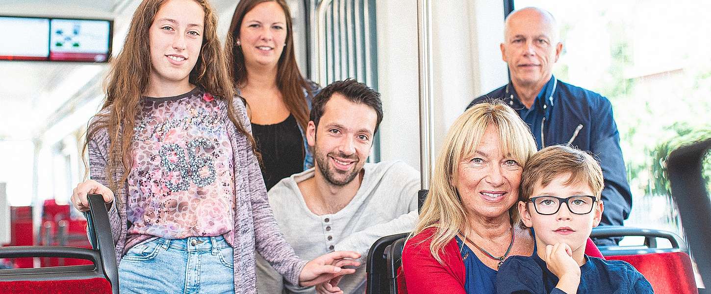 Eine Familie in der Bahn. Links ein brunettes Mädchen mit einer kurzen Jeans Shorts. Im Hintergrund lächelt eine brunette Dame in die Kamera. In der Mitte ein sitzt ein junger Mann umgedreht auf dem Sitz. Oma hat vorne rechts ihren Enkel auf dem Schoß.