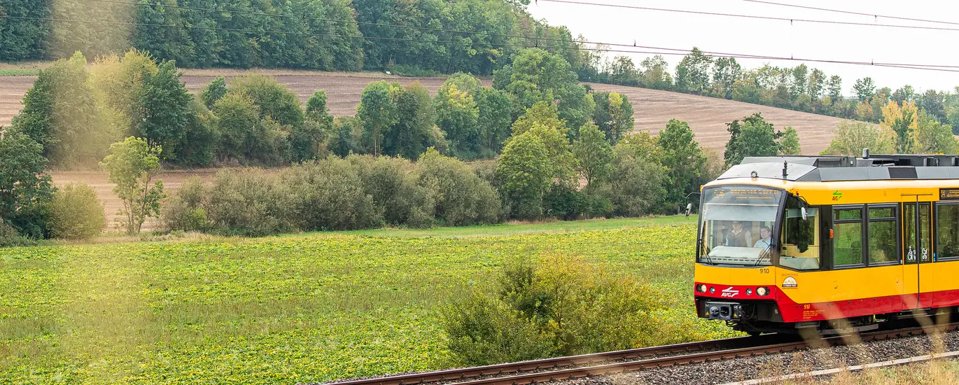 Eine AVG-Stadtbahn fährt auf einem eingleisigen Streckenabschnitt der Kraichgaubahn. Im Hintergrund sind Wiesen und bewaldete Hügel zu sehen.