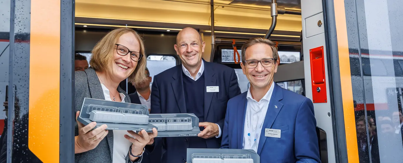 Das Foto zeigt drei Personen in der geöffneten Tür einer Tram-Train-Bahn. Abgebildet sind von links nach rechts Sonja Wiesholzer (Schiene Oberösterreich), Karsten Nagel (Saarbahn) und Christian Höglmeier (Albtal-Verkehrs-Geselllschaft/Verkehrsbetriebe Karlsruhe). Alle halten ein graues Fahrzeugmodell einer Bahn in der Hand. 